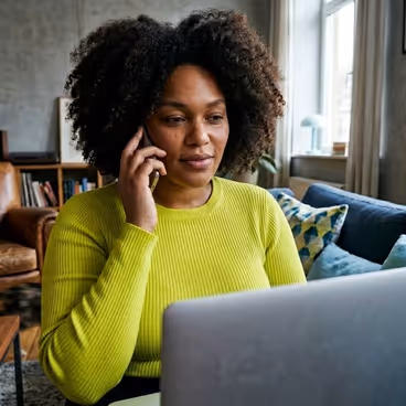 Een vrouw zit thuis achter de laptop terwijlze aan de telefoon is