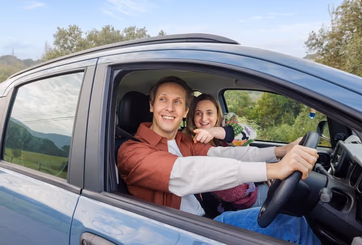 Een man en vrouw zitten i de auto en de vrouw wijst enthousiast uit het raam naar iets wat ze buiten ziet. (Alpaca's)