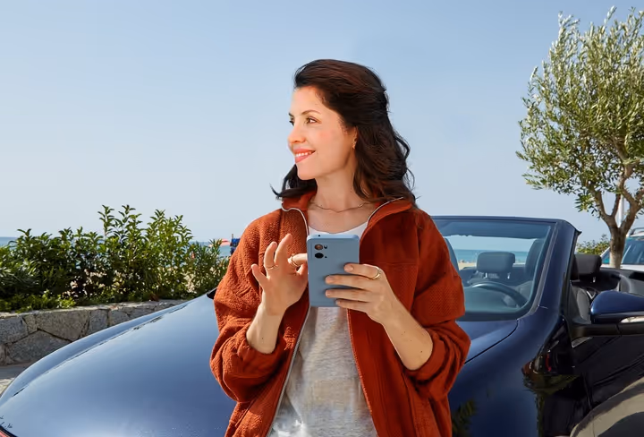 Een vrouw staat voor haar cabrio aan het strand en kijkt weg van de mobiel in haar hand naar opzij.