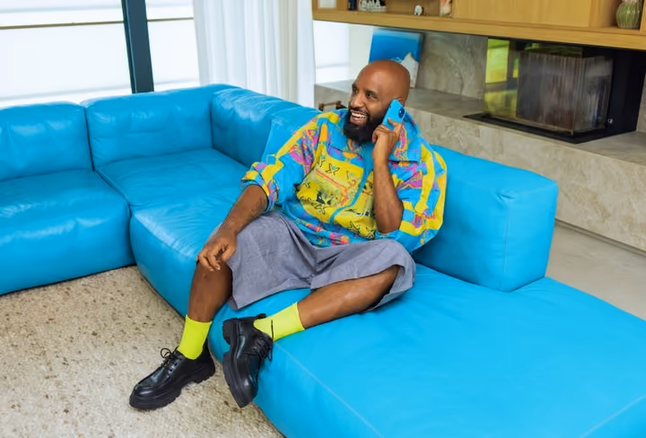 Willie Wartaal zit op een blauwe bank in een woonkamer met zijn mobiel in zijn hand en kijkt naar links