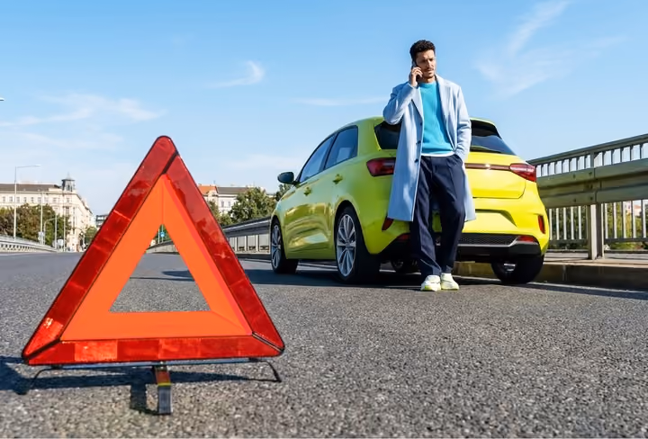 Een man zit in zijn auto die langs de weg staat met pech en een gevardendriehoek ernaast