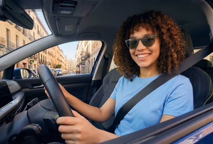 Een lachende vrouw zit achter het stuur van haar blauwe auto. 
