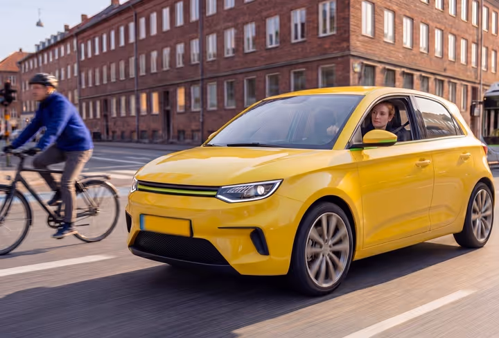 Een vrouw rijdt in een gele auto over een weg door de stad, met voor haar een fietser en achter haar een bus. 
