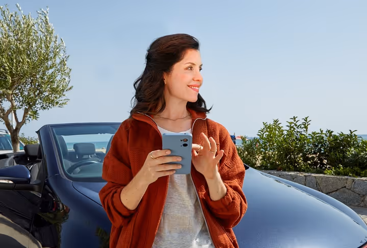 Een vrouw staat voor haar cabrio aan het strand en kijkt weg van de mobiel in haar hand naar opzij.