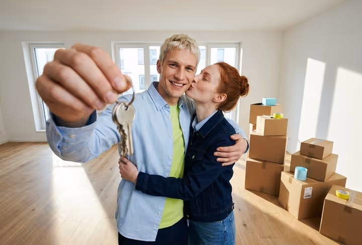 Een stel staat in een lege woonkamer en houdt trots de sleutel van de nieuwe woning in hun hand. 