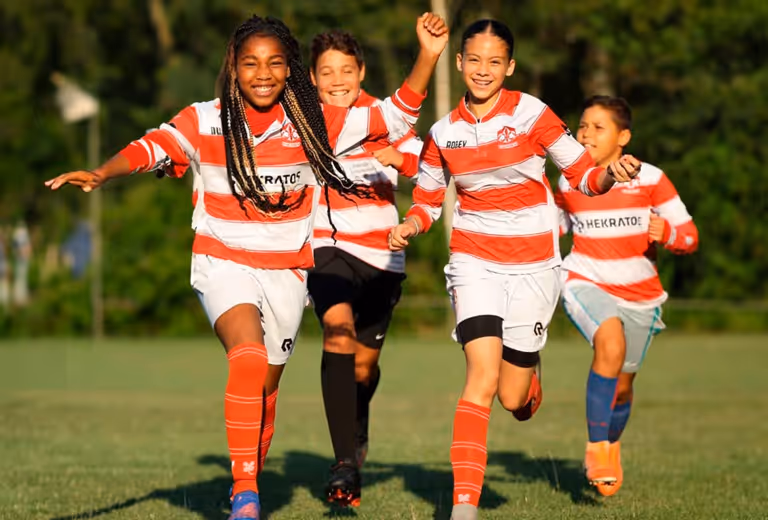 Vier kinderen in sporttenue rennen vrolijk over een sportveld richting de camera