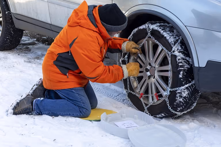 Man zit op z'n knieën in de sneeuw naast een auto om de sneeuwkettingen om de banden te doen.