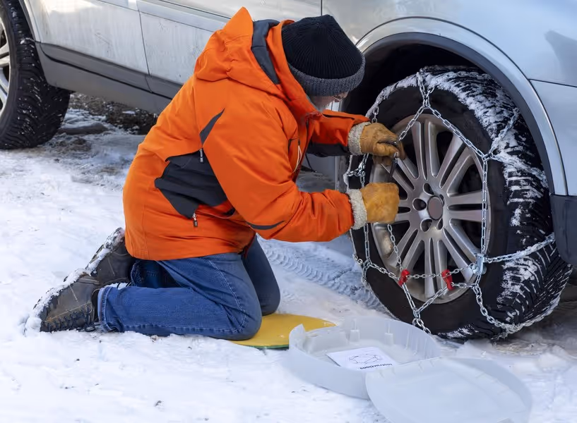 Man zit op z'n knieën in de sneeuw naast een auto om de sneeuwkettingen om de banden te doen. 
