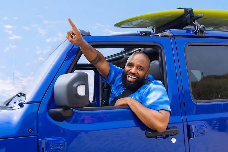 Willie Wartaal kijkt lachend uit zijn autoraam en wijst naar voren.
