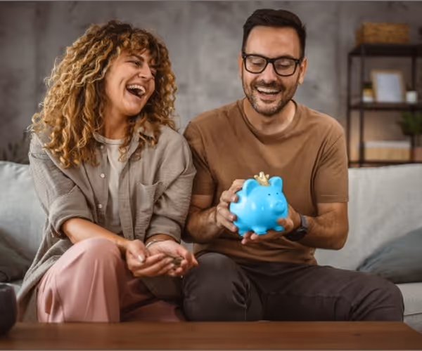 Een man en vrouw zitten lachend op de bank met een blauw spaarvarken in hun handen
