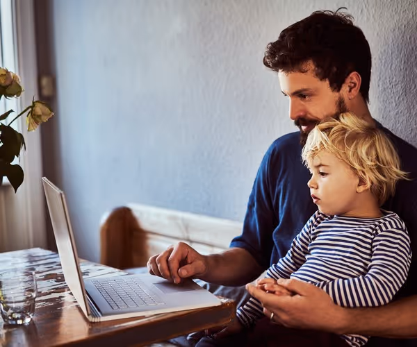 Een vader zit met een kindje op schoot achter zijn laptop