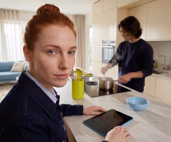 Eenvroiuw zit in de keuken waar een man nmet het eten bezig is en kijkt in de camera