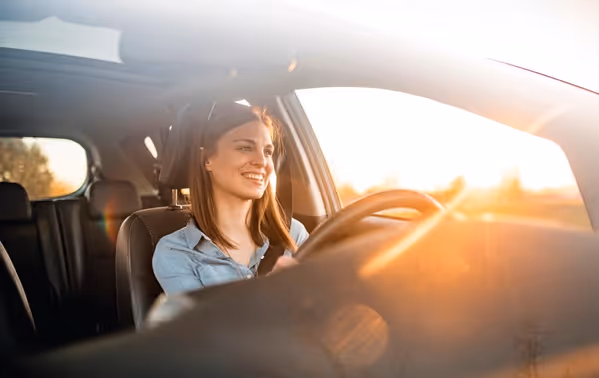 Een vrouw zit vrolijk achter het stuur van een auto terwijl de zon naar binnen schijnt